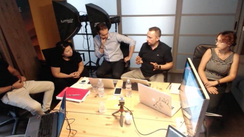 Six people sit around a wooden table in a studio, laptops open, talking and brainstorming a project.